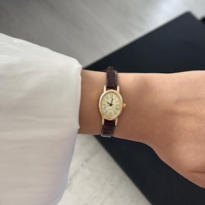May include: A gold-toned oval wristwatch with a brown leather band is worn on a wrist. The watch face has Roman numerals and the word "Quartz". The watch is on a wrist with a white shirt sleeve.
