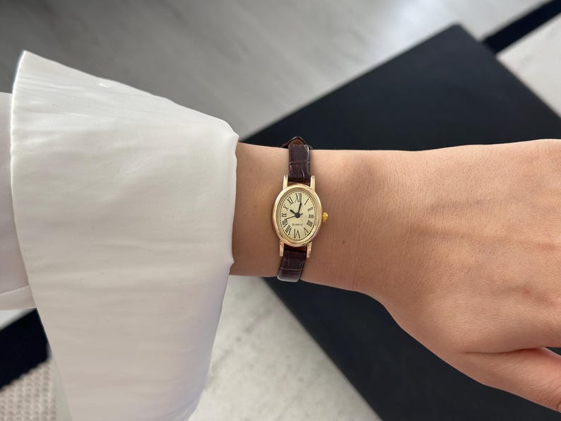 May include: A gold-toned oval wristwatch with a brown leather band is worn on a wrist. The watch face has Roman numerals and the word "Quartz". The watch is on a wrist with a white shirt sleeve.