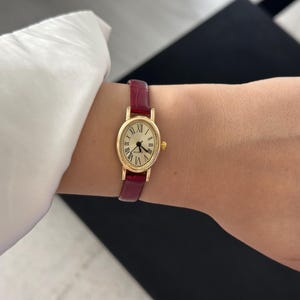 May include: A gold-toned wristwatch with a burgundy leather band. The watch face is oval-shaped with Roman numeral hour markers and black hands. The band is approximately 0.5 inches wide.