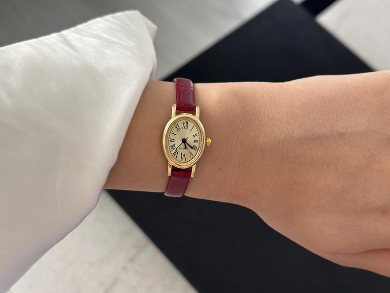 May include: A gold-toned wristwatch with a burgundy leather band. The watch face is oval-shaped with Roman numeral hour markers and black hands. The band is approximately 0.5 inches wide.