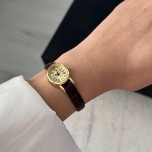 May include: A gold-toned wristwatch with a round face and Roman numeral hour markers. The watch has a dark brown leather band. The watch is on a wrist, and the background is blurred.