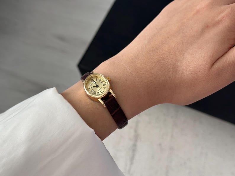 May include: A gold-toned wristwatch with a round face and Roman numeral hour markers. The watch has a dark brown leather band. The watch is on a wrist, and the background is blurred.