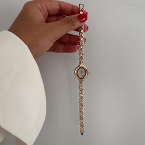 Op de afbeelding: Een goudkleurig polshorloge met een ovale wijzerplaat en een schakelband. Het horloge wordt tegen een witte achtergrond gehouden. De band heeft een reeks herhalende ovale en cirkelvormige schakels. De wijzerplaat heeft een lichtgeleurde achtergrond.