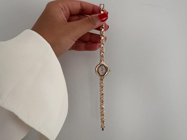 Op de afbeelding: Een goudkleurig polshorloge met een ovale wijzerplaat en een schakelband. Het horloge wordt tegen een witte achtergrond gehouden. De band heeft een reeks herhalende ovale en cirkelvormige schakels. De wijzerplaat heeft een lichtgeleurde achtergrond.