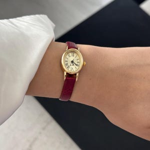 May include: A gold-toned oval wristwatch with a cream-colored face and black Roman numerals. The watch has a burgundy leather band. The watch is on a wrist, next to a white long-sleeved shirt.