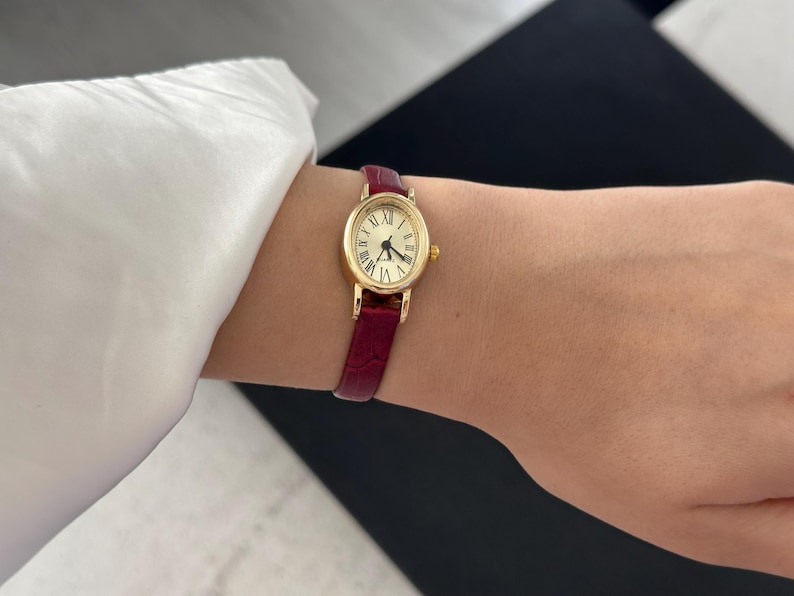 May include: A gold-toned oval wristwatch with a cream-colored face and black Roman numerals. The watch has a burgundy leather band. The watch is on a wrist, next to a white long-sleeved shirt.