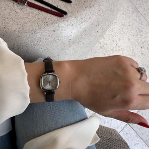 May include: A silver wristwatch with a square face and a dark brown leather strap. The watch is worn on a wrist, with a matching dark brown watch strap visible in the background. The watch face has a light silver colour.