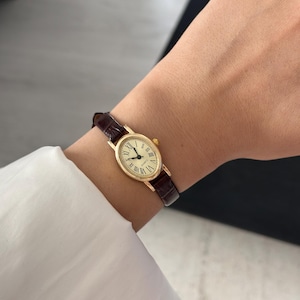 May include: A gold-toned wristwatch with a brown leather band. The watch face is oval with Roman numerals and black hands. The watch is worn on a wrist, with a white shirt sleeve visible in the background.