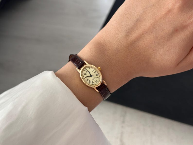 May include: A gold-toned wristwatch with a brown leather band. The watch face is oval with Roman numerals and black hands. The watch is worn on a wrist, with a white shirt sleeve visible in the background.