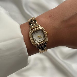 May include: A gold-tone quartz wristwatch featuring a square-shaped case with rounded edges and a white dial. The watch face displays black Roman numeral hour markers and thin gold hands. It includes a metallic link bracelet with a repeating pattern.