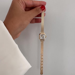 Op de afbeelding: Een goudkleurig polshorloge met een vierkante wijzerplaat en een mesh band. Het horloge heeft een lichtgekleurde wijzerplaat en is versierd met kleine, ronde versieringen langs de band. Het horloge wordt tegen een effen achtergrond gehouden.