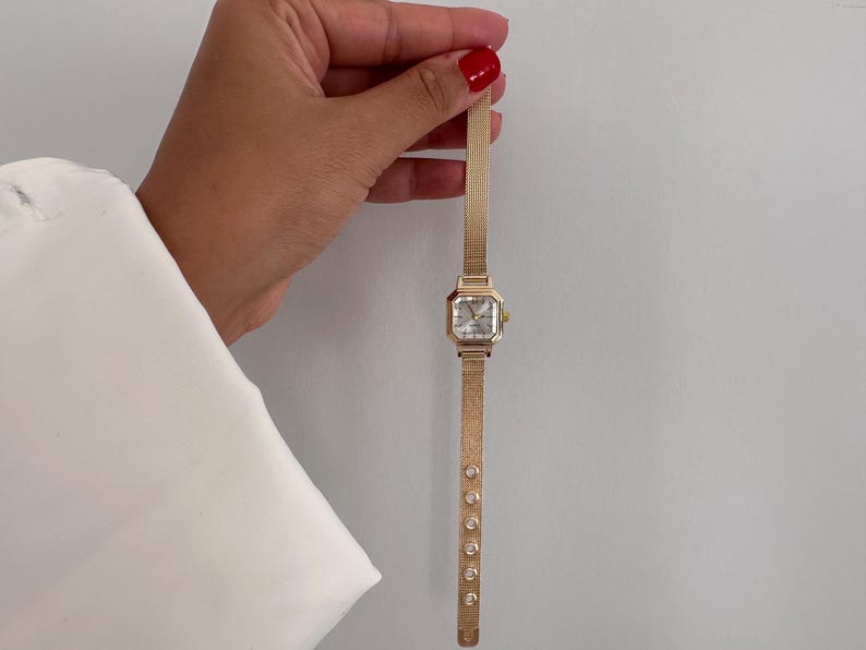 Op de afbeelding: Een goudkleurig polshorloge met een vierkante wijzerplaat en een mesh band. Het horloge heeft een lichtgekleurde wijzerplaat en is versierd met kleine, ronde versieringen langs de band. Het horloge wordt tegen een effen achtergrond gehouden.