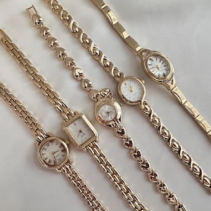 Può includere: Cinque orologi da polso color oro con vari design di cinturini e quadranti. Gli orologi hanno quadranti rotondi e quadrati con numeri romani. I cinturini presentano motivi a maglie, a cuore e decorativi. Gli orologi sono esposti su una superficie bianca.