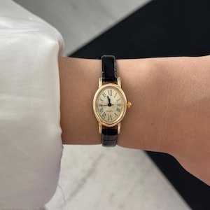 May include: A gold-toned quartz watch with a cream-colored face and black Roman numerals. The watch has a black leather band. The watch is oval-shaped and is worn on the wrist.