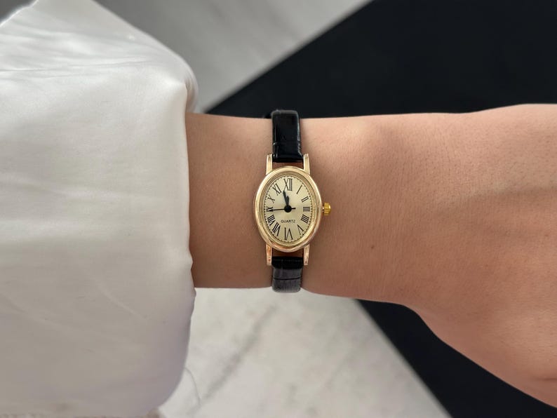 May include: A gold-toned quartz watch with a cream-colored face and black Roman numerals. The watch has a black leather band. The watch is oval-shaped and is worn on the wrist.