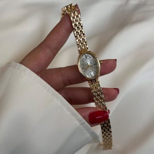 Könnte beinhalten: Goldfarbene Armbanduhr mit ovalem Zifferblatt und Gliederarmband. Das Zifferblatt ist silberfarben mit goldenen Zeigern und römischen Ziffern. Das Armband hat eine passende Goldfarbe.