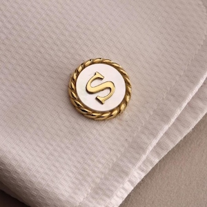 May include: A gold-toned cufflink with the letter "S" in the center, set against a white background. The cufflink is round with a rope-like border. The cufflink is resting on a white textured fabric.