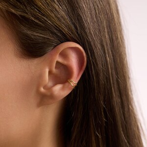 Puede incluir: Primer plano de una oreja con un ear cuff dorado. El ear cuff tiene un dise&ntilde;o de doble banda y se lleva en la parte superior de la oreja. La persona tiene el pelo largo y casta&ntilde;o.