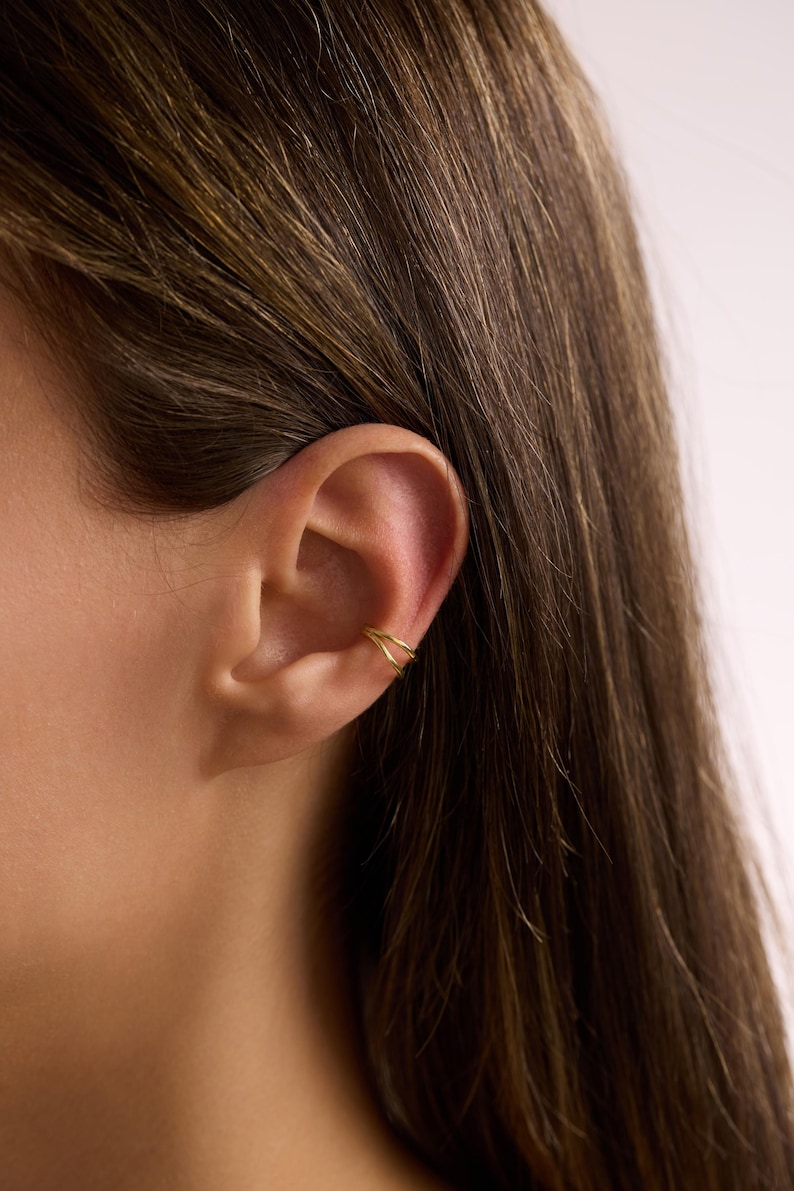 Puede incluir: Primer plano de una oreja con un ear cuff dorado. El ear cuff tiene un dise&ntilde;o de doble banda y se lleva en la parte superior de la oreja. La persona tiene el pelo largo y casta&ntilde;o.