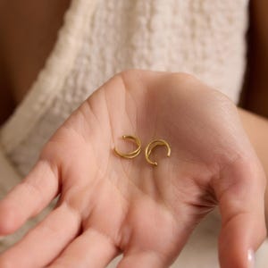 Puede incluir: Dos pendientes de oreja dorados se muestran en una palma abierta. Los pendientes tienen un dise&ntilde;o curvo y minimalista. El fondo est&aacute; borroso, con una tela de color crema visible.