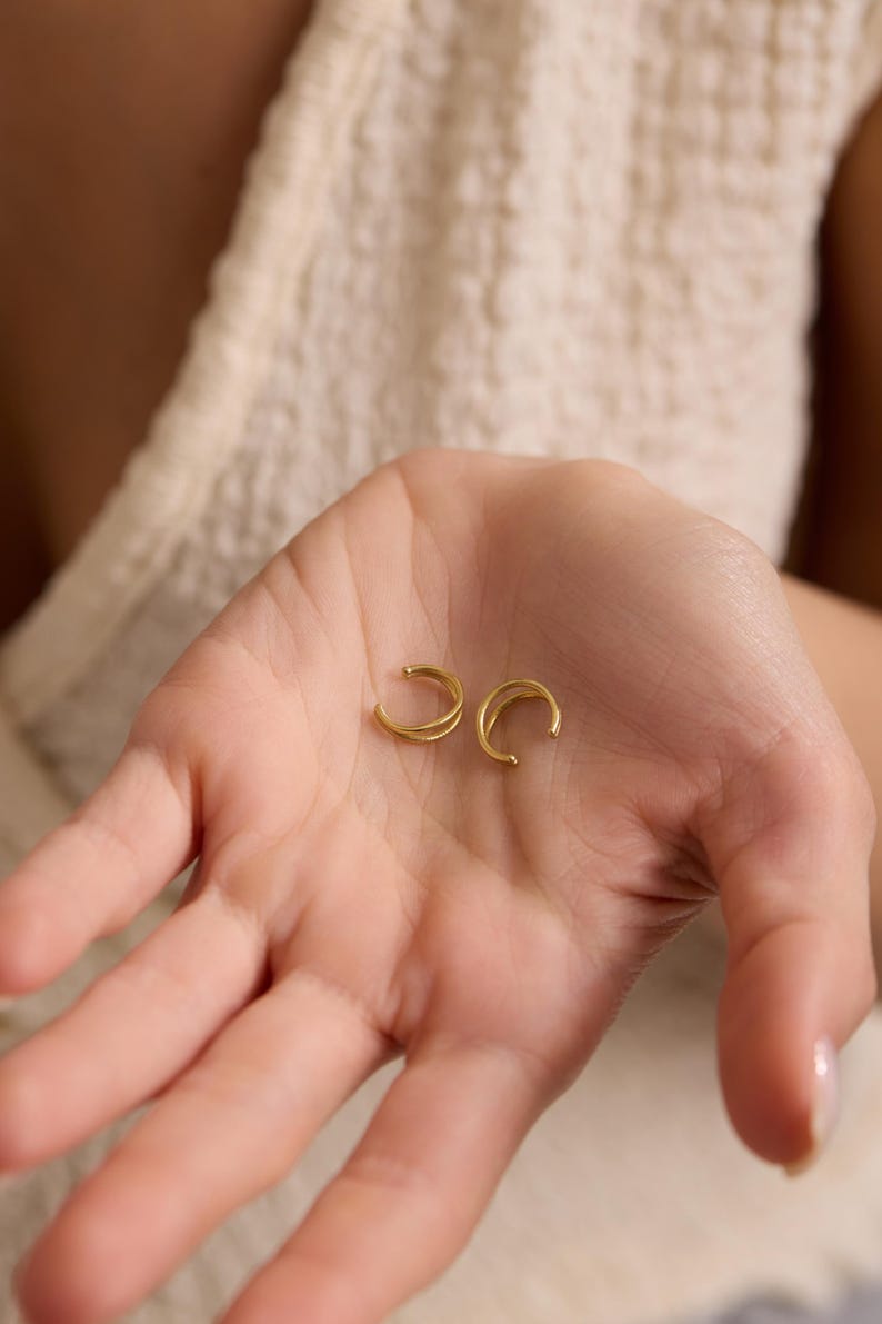 Puede incluir: Dos pendientes de oreja dorados se muestran en una palma abierta. Los pendientes tienen un dise&ntilde;o curvo y minimalista. El fondo est&aacute; borroso, con una tela de color crema visible.