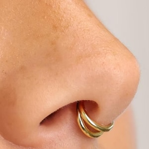 Puede incluir: Primer plano de una nariz con un aro de nariz dorado de doble aro. El aro de la nariz tiene un dise&ntilde;o sencillo y elegante. La piel alrededor de la nariz es suave y tiene un tono natural. La joya es un accesorio de moda.