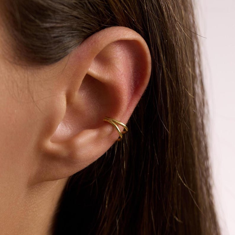Puede incluir: Un ear cuff dorado con un dise&ntilde;o de doble banda. El cuff se envuelve alrededor del cart&iacute;lago de la oreja, creando una joya minimalista y moderna. La joya se muestra en la oreja de una persona.