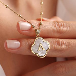 May include: Gold-toned necklace featuring a four-leaf clover pendant with a mother-of-pearl inlay and the letter "A". The necklace has a delicate chain with small, round beads. The pendant is held by a hand with manicured nails.