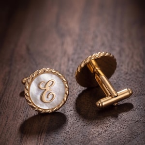 May include: A pair of gold-toned cufflinks featuring a circular design with a white mother-of-pearl inlay. Each cufflink is bordered by a twisted rope-style metal edge and displays a prominent, elegant gold-colored letter E in the center.