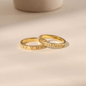 Puede incluir: Dos anillos de boda dorados. Un anillo tiene grabada la fecha en números romanos XX.V.MM. El otro anillo tiene grabados los nombres "Thomas & Jessica". Los anillos están sobre una superficie blanca.