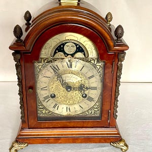 Puede incluir: Un reloj antiguo de madera con asa de latón y detalles decorativos. La esfera del reloj presenta números romanos y detalles ornamentados. El reloj tiene un indicador de fase lunar y está sostenido por patas de latón.