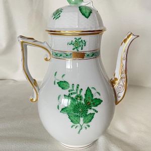 Puede incluir: Cafetera de porcelana blanca con tapa, asa y boquilla. Decorada con diseños florales verdes y ribetes dorados. La tapa presenta un asa en forma de hoja verde. Una pieza clásica para servir café o té.