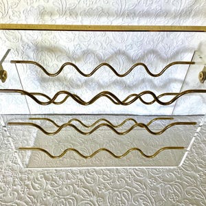 May include: A clear acrylic wine rack with a gold-colored metal frame and wavy supports. The rack is designed to hold multiple bottles and has a rectangular shape with a decorative top bar. The overall design is modern and elegant.