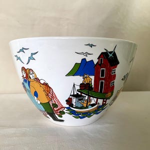 Op de afbeelding: Een witte keramische kom met een kleurrijke illustratie van een kusttafereel. Het ontwerp bevat figuren in gele en blauwe kleding die een visnet vasthouden, een rood huis op palen, een kleine boot op blauwe golven en vliegende meeuwen.