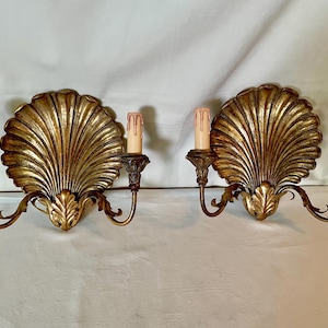 Op de afbeelding: Een paar goudkleurige wandlampen, elk met een schelpvormige achterplaat met stralende ribbels. Twee gebogen armen steken uit de basis en ondersteunen kaarsvormige armaturen met bleke, druipende afdekkingen aan weerszijden.
