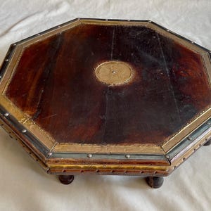 Può includere: Tavolo ottagonale in legno con superficie marrone scuro e finiture in ottone. Il tavolo ha piccole gambe arrotondate e un medaglione decorativo in ottone al centro. I bordi sono bordati con piccoli borchie metalliche.
