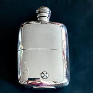 Könnte beinhalten: Ein polierter, silberfarbener Metallflachmann mit abgerundeter rechteckiger Form und Schraubverschluss. Die Vorderseite zeigt ein kleines, mittig platziertes Kreuzemblem nahe der Unterkante. Das Metall hat eine reflektierende Oberfläche.