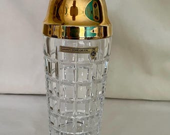 May include: A cocktail shaker with a clear, grid-patterned glass body and a polished gold-tone metal lid. A rectangular label on the glass reads GARANTI DORE OR FIN with a small circular logo featuring the letters LC.