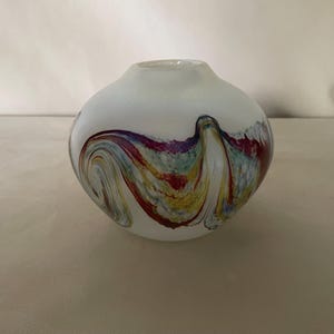 Può includere: Un vaso sferico in vetro con una base bianca opaca caratterizzato da un motivo vorticoso multicolore nei toni del rosso, giallo, blu e viola. Il vaso presenta una piccola apertura circolare sulla parte superiore e un corpo liscio e rotondo.