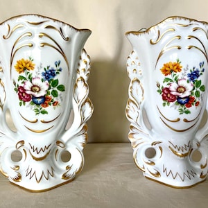 May include: A pair of white porcelain wall pockets featuring a central floral bouquet with yellow, pink, red, and blue flowers. The vessels have ornate, scalloped edges and relief details accented with gold trim, including two circular cutouts at the base.