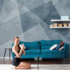 Può includere: Una donna in abbigliamento sportivo nero si inginocchia su un tappetino da yoga, con le mani giunte in preghiera. Uno smartphone su un treppiede registra la scena. Un divano turchese, arte murale geometrica e un tavolino completano l'ambiente.