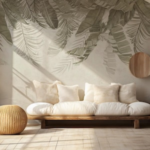 May include: A light-filled living room with a wooden sofa, white cushions, and a woven ottoman. The wall features a large mural of green and gray palm leaves. A white vase holds dried plants.