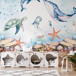 Op de afbeelding: Een grillige kinderkamerscène met een witte opbergunit met uitgesneden vormen van een ster en walvissen. De achtergrond is een aquarelmuurschildering van het zeeleven, waaronder walvissen, een zeeschildpad en zeesterren.