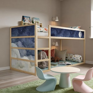 Op de afbeelding: Een hoogslaper van lichtgekleurd hout met zijpanelen voorzien van een donkerblauwe nachthemelprint, sterren en een maan. Het onderste gedeelte bevat een grijze pluche olifant, kussens en een kussenrol. Een ronde witte tafel met lichtblauwe en roze stoelen staat in de buurt.