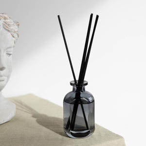May include: A gray glass aromatherapy diffuser bottle with black reeds. The bottle has a rounded shape and contains a clear liquid. The reeds are inserted into the bottle, and the background is a neutral color.