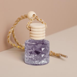 May include: A small, clear glass bottle with a geometric pattern, filled with a lavender-colored liquid. It has a wooden cap and a braided rope for hanging. The rope is beige with gold accents. The bottle is designed for aromatherapy or fragrance.