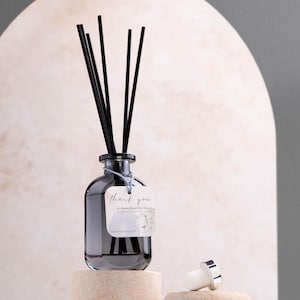 Puede incluir: Un difusor de vidrio gris oscuro con varillas negras y una etiqueta blanca que dice "thank you". La botella está sobre un bloque beige, con un tapón plateado y blanco cerca. Es un artículo de fragancia para el hogar.