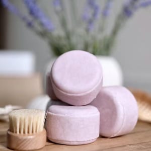 May include: A collection of light purple, round solid shampoo bars are stacked on a wooden surface. A small wooden brush with white bristles sits in front of the bars. A vase of lavender flowers is in the background.