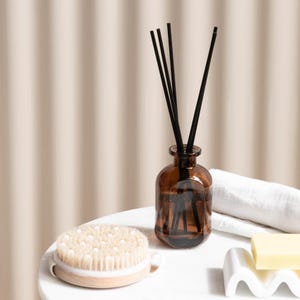 Puede incluir: Un frasco difusor de vidrio marrón con cañas negras, un cepillo corporal con mango de madera, una toalla blanca y una pastilla de jabón sobre una superficie blanca. Los artículos crean un ambiente de spa.