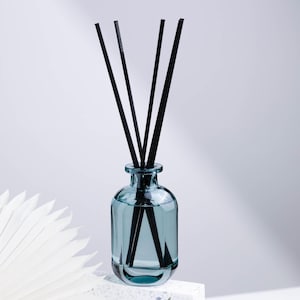 Puede incluir: Un difusor de vidrio color turquesa con varillas negras. La botella contiene un líquido transparente y está colocada sobre una superficie blanca. Las varillas están dispuestas en forma de abanico, creando una estética minimalista.